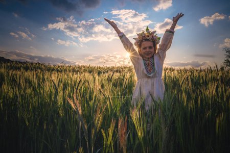 Téléchargez les photos : Belle jeune fille avec chapelet de fleurs, robe folklorique ethnique avec broderie bulgare traditionnelle au coucher du soleil sur un champ agricole de blé - en image libre de droit