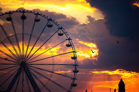 Photo for Ferris Wheel and illuminations in amusement park during scenic sunset with dramatic sky clouds and flying birds - Royalty Free Image