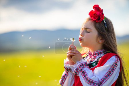 Photo for Little girl blowing dandelion on a green field. Bulgarian woman in ethnic folklore dress blow dandelions. - Royalty Free Image