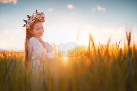 Téléchargez les photos : Belle jeune fille avec chapelet de fleurs, robe folklorique ethnique avec broderie bulgare traditionnelle au coucher du soleil sur un champ agricole de blé - en image libre de droit