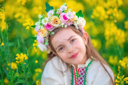 Foto de Soñando Hermosa joven con chaplet de flores, vestido de folclore étnico con bordado búlgaro tradicional durante la puesta del sol en un campo agrícola de semillas de colza - Imagen libre de derechos