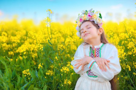 Téléchargez les photos : Rêver Belle jeune fille avec chapelet de fleurs, robe folklorique ethnique avec broderie bulgare traditionnelle au coucher du soleil sur un champ agricole de colza - en image libre de droit