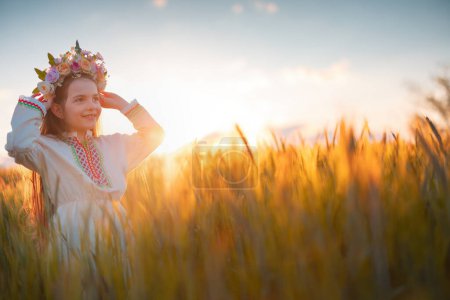 Foto de Hermosa joven con chaplet de flores, vestido de folclore étnico con bordado búlgaro tradicional durante la puesta del sol en un campo agrícola de trigo - Imagen libre de derechos