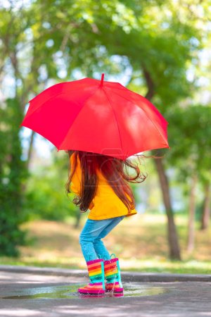 Foto de Feliz chica hermosa con paraguas y botas de goma de color saltar en un charco lluvioso - Imagen libre de derechos