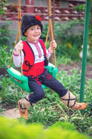 Téléchargez les photos : Garçon bulgare en costume folklorique ethnique avec broderie traditionnelle sur une balançoire dans un jardin de sa maison familiale - en image libre de droit