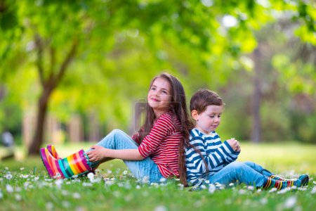 Foto de Feliz niño y niña en botas de goma de lluvia jugando al aire libre en el parque verde con el campo floreciente de flores de margarita - Imagen libre de derechos
