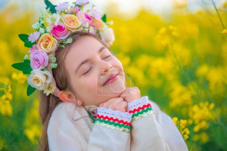 Téléchargez les photos : Champ de colza et fille rêveuse avec chapelet de fleurs, robe folklorique ethnique avec broderie bulgare traditionnelle - en image libre de droit