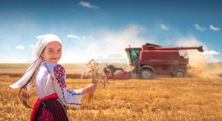 Photo for Girl in traditional ethnic folklore costume with Bulgarian embroidery standing on a harvest golden wheat fiel with industrial combine machine - Royalty Free Image