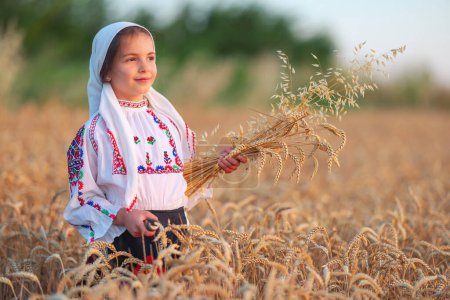 Téléchargez les photos : Champ de récolte de blé et agricultrice bulgare petite fille en costume folklorique ethnique tenir pailles de blé doré et la faucille, la récolte et l'agriculture en Bulgarie - en image libre de droit
