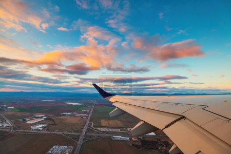 Photo for Airplane flight in sunset sky over city and wing of plane. View from the window of the Aircraft. Traveling in air. - Royalty Free Image