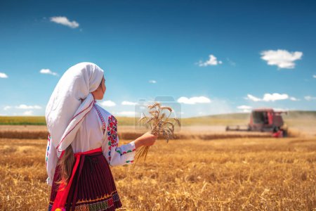 Foto de Chica joven con traje tradicional de folclore búlgaro en el campo de trigo agrícola durante el tiempo de cosecha con máquina combinadora industrial - Imagen libre de derechos