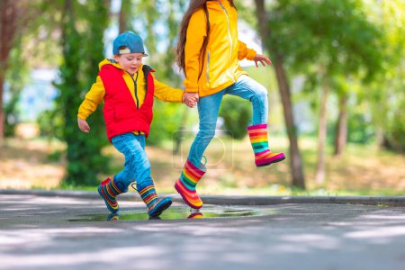 Téléchargez les photos : Enfants heureux fille et garçon avec parapluie et bottes de pluie en caoutchouc coloré jouer en plein air et sauter dans la flaque d'eau pluvieuse - en image libre de droit