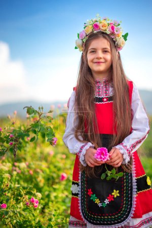Foto de Chica búlgara con corona de flores recogiendo aceite que lleva rosas damascena en un campo de rosas en flor al amanecer - Imagen libre de derechos