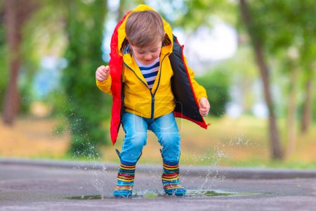 Foto de Niño feliz con paraguas y botas de goma de color saltar en un charco lluvioso en un parque, infancia despreocupada - Imagen libre de derechos