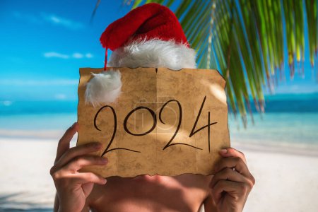 Téléchargez les photos : Vacances saisonnières d'hiver sur une île tropicale pour la nouvelle année 2024, homme avec chapeau de Père Noël et trésor vieille carte sur une plage exotique avec des palmiers - en image libre de droit