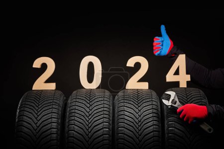 Foto de Servicio de neumáticos de coche de invierno y pulgares manos arriba de mecánico y texto 2024 feliz año nuevo sobre fondo negro - Imagen libre de derechos