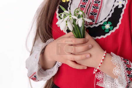 Photo for Bulgarian girl in ethnic folklore embroidery dress nosia, yarn bracelet martenitsa, and spring bouquet snowdrops symbol of Bulgaria - Royalty Free Image