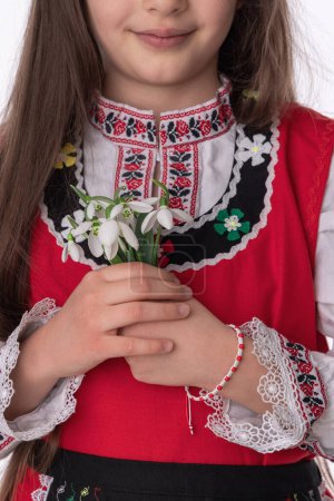 Foto de Chica búlgara en trajes folclóricos tradicionales con flores de primavera ramo de gotas de nieve y artesanía pulsera de lana símbolo martenitsa de Baba Marta vacaciones, primavera y marzo mes - Imagen libre de derechos