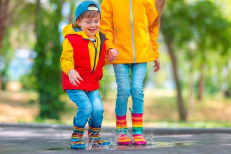 Foto de Feliz niña y niño con paraguas y botas de goma color lluvia jugar al aire libre y saltar en charcos lluviosos sucios - Imagen libre de derechos