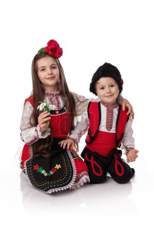 Photo for Bulgarian girl and boy in ethnic folklore costumes with spring traditional martenitsa on hands and bouquet of snowdrops, happy Baba Marta day, Bulgaria - Royalty Free Image