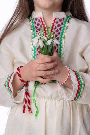 Bulgarian girl in ethnic costume with bouquet of snowdrops and a red and white symbol of the holiday of the spring martenitsa Baba Marta day, Bulgaria