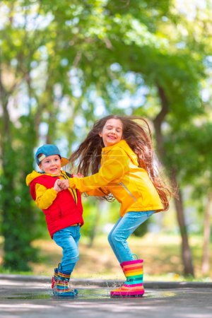 Foto de Chica y niño feliz con botas de lluvia de goma coloridas jugando al aire libre y saltando en charco lluvioso - Imagen libre de derechos