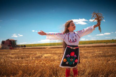 Foto de Ropa de niña búlgara traje de bordado folclore en el campo de trigo agrícola durante la cosecha y combinar la máquina - Imagen libre de derechos