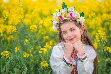 Dreaming beautiful young girl with spring flower chaplet, ethnic folklore dress with traditional bulgarian embroidery on a yellow rapeseed field