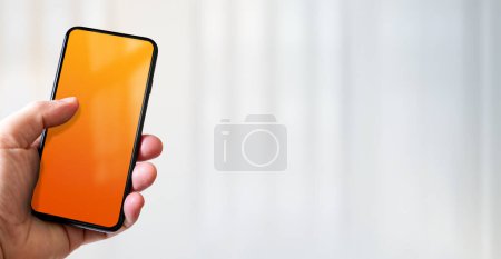 Photo for Hand holding a smartphone with blank orange screen. White office background. Horizontal banner. - Royalty Free Image
