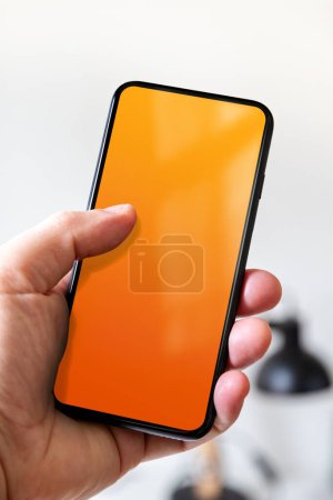 Photo for Hand holding a smartphone with blank orange screen. White office background. - Royalty Free Image