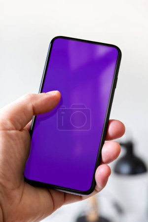 Photo for Hand holding a smartphone with blank purple screen. White office background. - Royalty Free Image