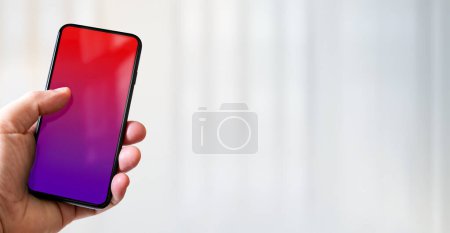 Foto de Mano sosteniendo un teléfono inteligente con pantalla roja y púrpura en blanco. Fondo de la oficina blanca. Banner horizontal. - Imagen libre de derechos