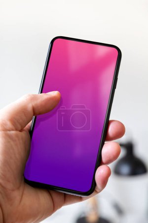 Photo for Hand holding a smartphone with blank pink and purple screen. White office background. - Royalty Free Image