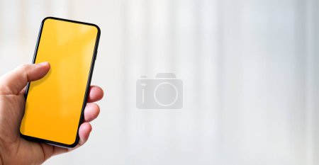 Photo for Hand holding a smartphone with blank yellow screen. White office background. Horizontal banner. - Royalty Free Image