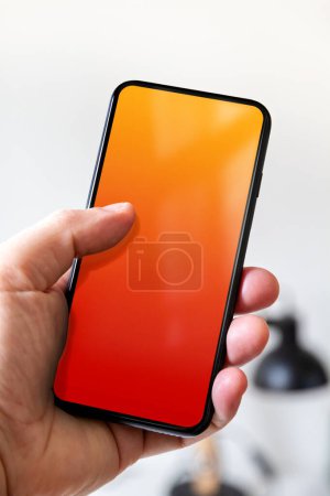 Téléchargez les photos : Hand holding a smartphone with blank red and orange screen. White office background. - en image libre de droit