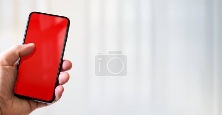 Photo for Hand holding a smartphone with blank red screen. White office background. Horizontal banner. - Royalty Free Image