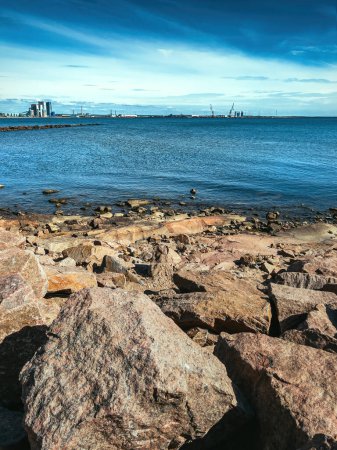 Photo for Rocky shoreline and Kattegat sea with Halmstad city industrial seaport in background on sunny summer day - Royalty Free Image
