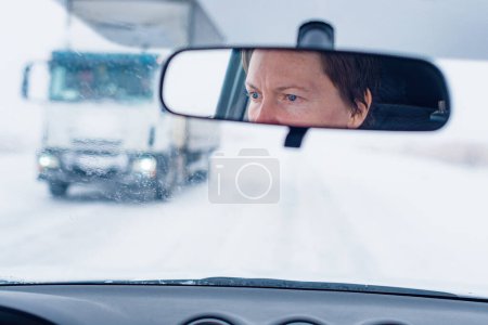 Photo for Face of a female driver in car rear-view mirror while driving in bad conditions during snow blizzard, selective focus - Royalty Free Image
