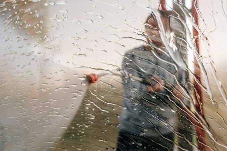 Téléchargez les photos : Man washing car with water gun in carwash self-service. Soap sud, wax and water drops covering vehicle window glass. Seen from the inside of the automobile. - en image libre de droit