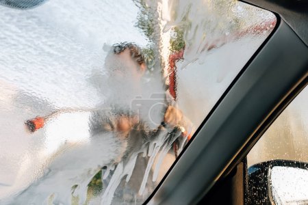 Foto de Man washing car with water gun in carwash self-service. Soap sud, wax and water drops covering vehicle window glass. Seen from the inside of the automobile. - Imagen libre de derechos