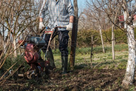 Foto de Agricultor que realiza labranza del suelo del jardín con la vieja máquina agrícola pobre del cultivador del cultivador, foco selectivo - Imagen libre de derechos