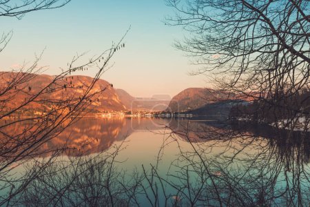 Téléchargez les photos : Belle image panoramique du coucher du soleil du lac Bohinj et du vieux village slovène Stara Fuzina en arrière-plan vu à travers des branches d'arbres à feuilles caduques au bord de l'eau en février - en image libre de droit
