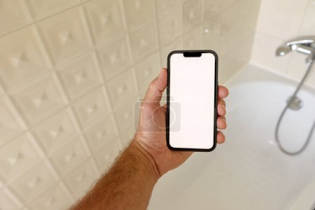 Téléchargez les photos : Modèle de smartphone dans la salle de bain, homme tenant un téléphone portable avec écran tactile vierge sur la baignoire, mise au point sélective - en image libre de droit