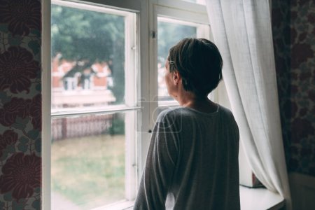 Téléchargez les photos : Initiation d'une femme moyenne adulte regardant par la fenêtre et réfléchissant à une orientation sélective - en image libre de droit