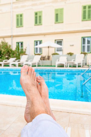 Photo for Barefoot male tourist relaxing by the outdoor swimming pool in seaside resort on sunny summer day, pov shot with selective focus - Royalty Free Image