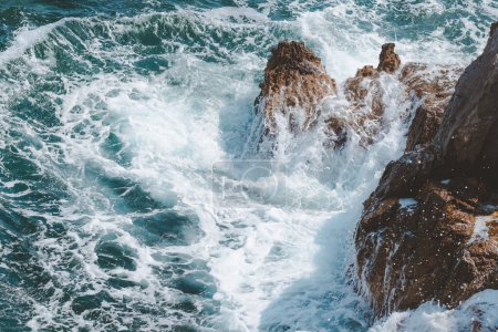 Foto de Ondas oceánicas frenando en las rocas de la costa cubriéndolas de espuma blanca en el soleado día de verano - Imagen libre de derechos