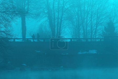 Foto de Dos personas caminando sobre el viejo puente de piedra en el lago Bohinj en la nebulosa mañana de invierno, imagen tonificada azul - Imagen libre de derechos