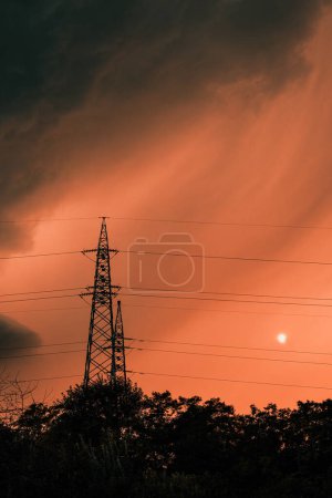 Téléchargez les photos : Nuages orageux sombres et menaçants au ciel orangé d'été, silhouettes de cimes d'arbres et pylône électrique juste avant la pluie, mise au point sélective - en image libre de droit