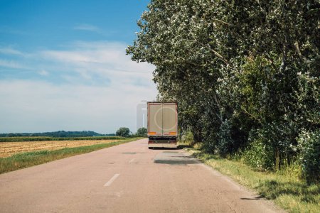 Photo for Trucking and freight transportation, rear view of large semi-truck driving along the road through countryside landscape in summer, diminishing perspective - Royalty Free Image