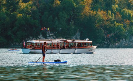 Téléchargez les photos : Bohinj, Slovénie - 27 août 2023 : L'homme sur la planche à pagaie par le bateau de tourisme électrique sur le lac Bohinj dans le parc national de Triglav - en image libre de droit